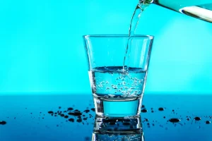 Klares Wasser wird in ein kurzes Glas gegossen, das auf einer glänzenden Oberfläche steht.