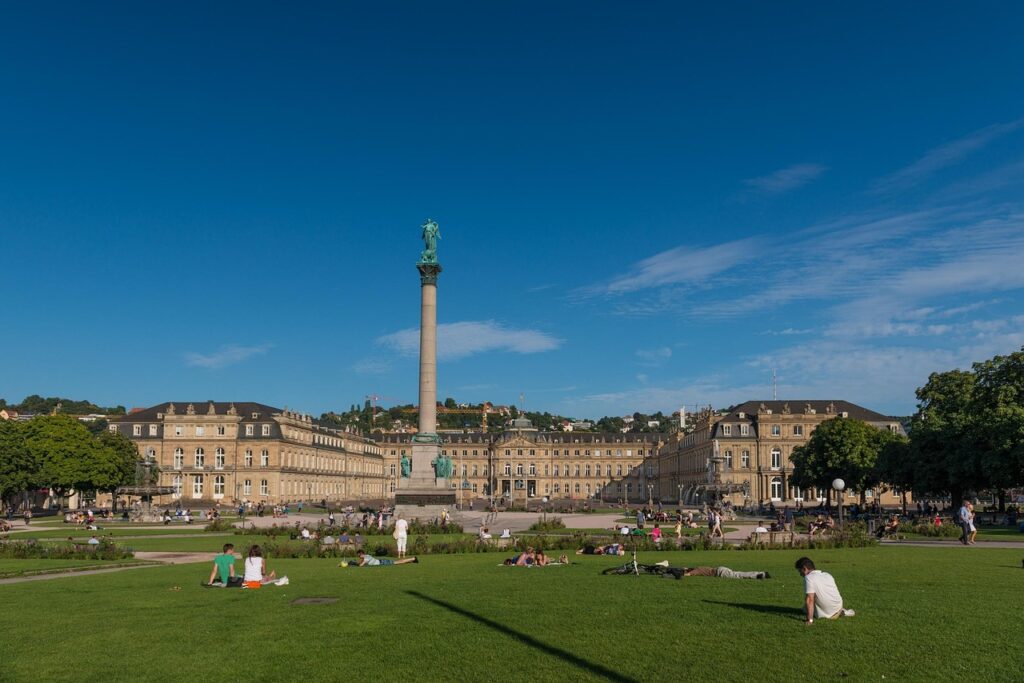 Panoramaansicht des Schlossplatzes in Stuttgart mit dem historischen Schloss und der Säule im Vordergrund, ideal für IT-Service Stuttgart & Umgebung. Menschen entspannen auf der Wiese unter blauem Himmel.