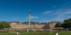 Panoramaansicht des Schlossplatzes in Stuttgart mit dem historischen Schloss und der Säule im Vordergrund, ideal für IT-Service Stuttgart & Umgebung. Menschen entspannen auf der Wiese unter blauem Himmel.